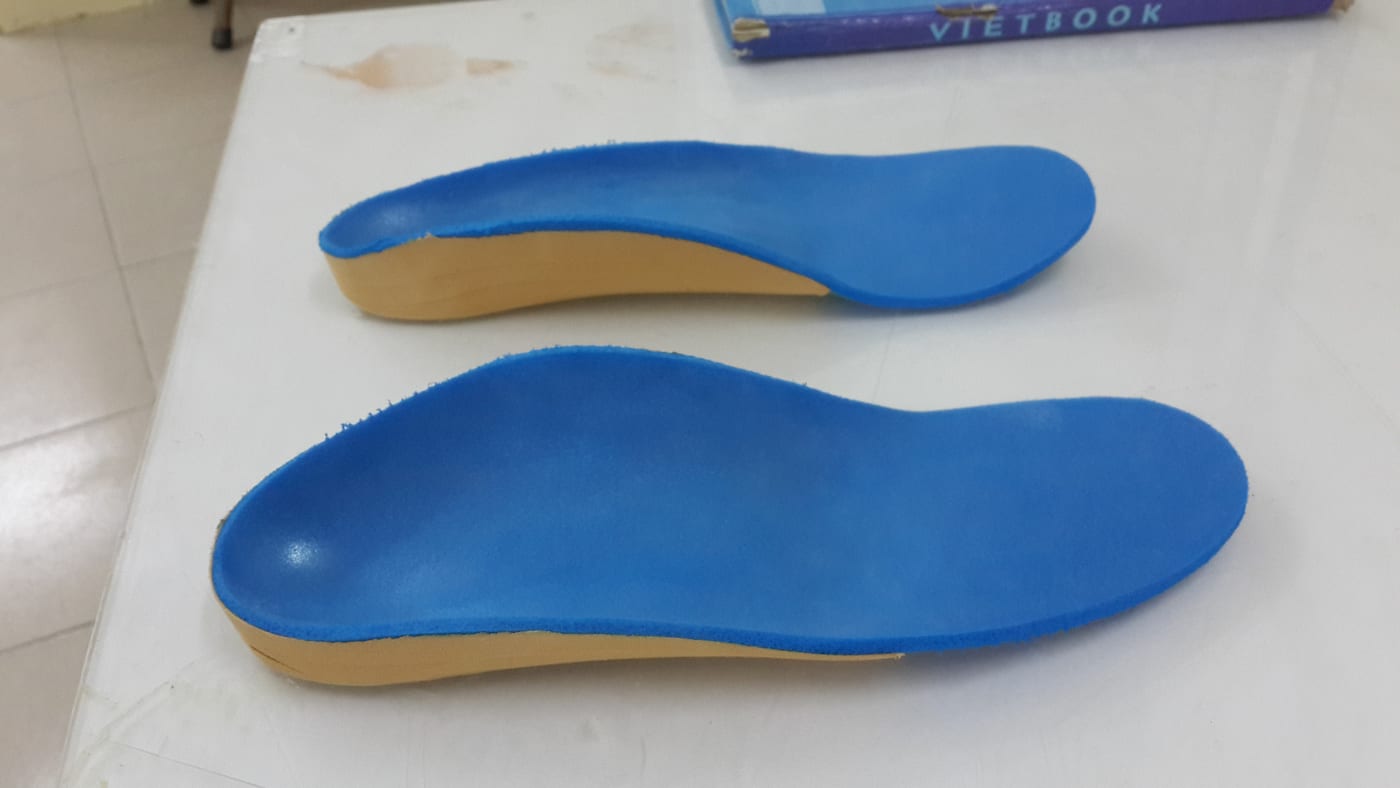 A custom insole (foot orthosis): the shell cradles the arch, a soft blue top layer contacts the foot, and a firmer base holds the shape. A metatarsal pad can be added inside to offload the painful space.